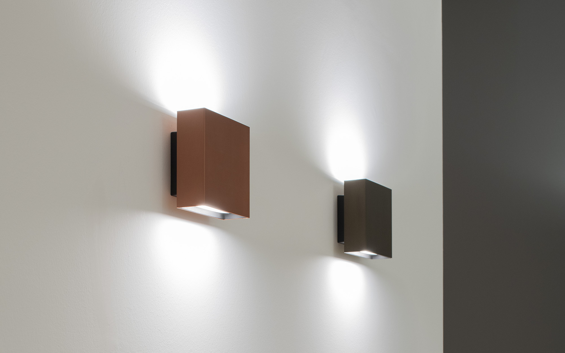 Due applique quadrate nelle finiture rame e bronzo scuro, che emettono fasci di luce verso l'alto e il basso su una parete bianca.