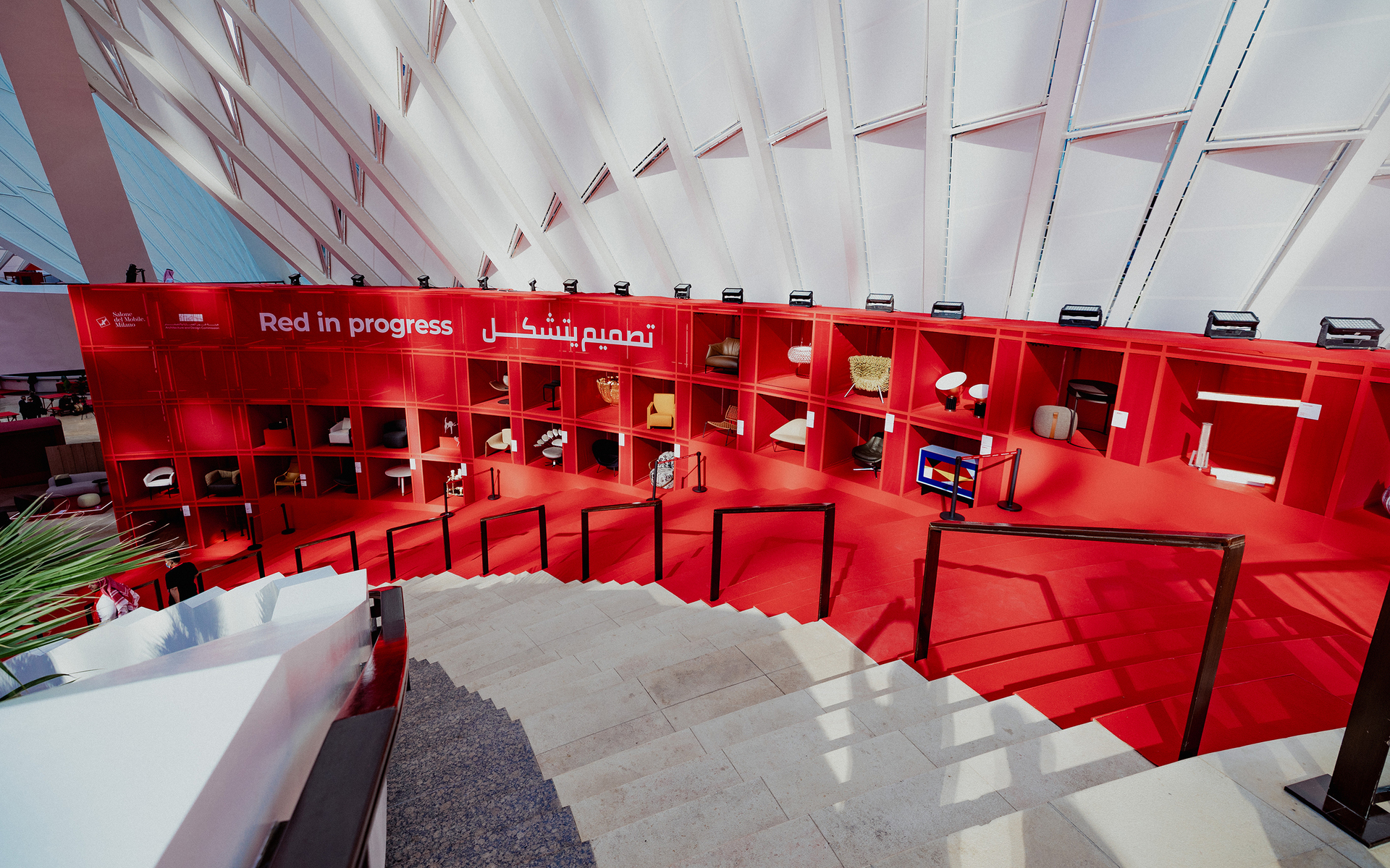 Mostra Red in Progress: parete espositiva rossa con oggetti di design e lampade su mensole, segnaletica in arabo e inglese, vista dalle scale.