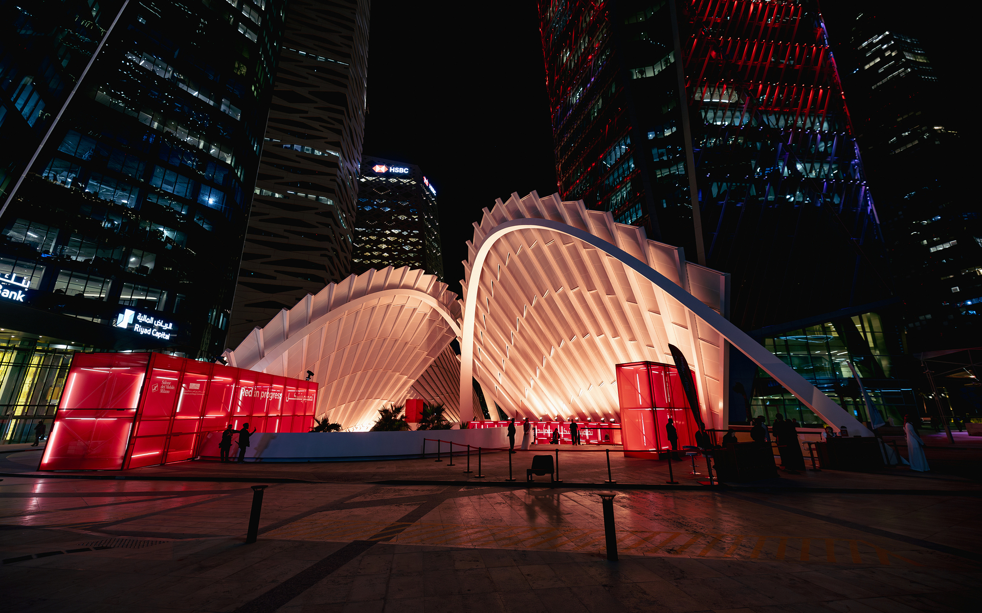 Padiglione architettonico bianco illuminato con copertura ad arco nervata e pannelli laterali a luce rossa, su sfondo di skyline notturno