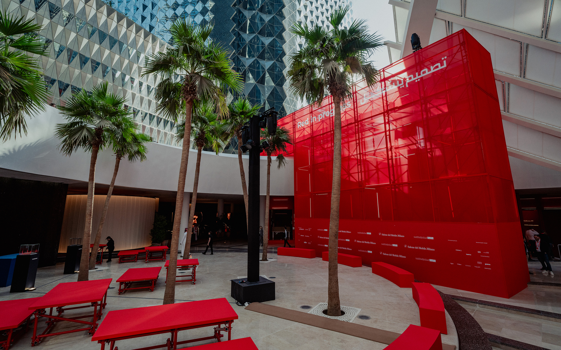 Piazza esterna con panchine rosse, palme e una grande struttura traslucida rossa con scritta araba, affiancata da edifici moderni.