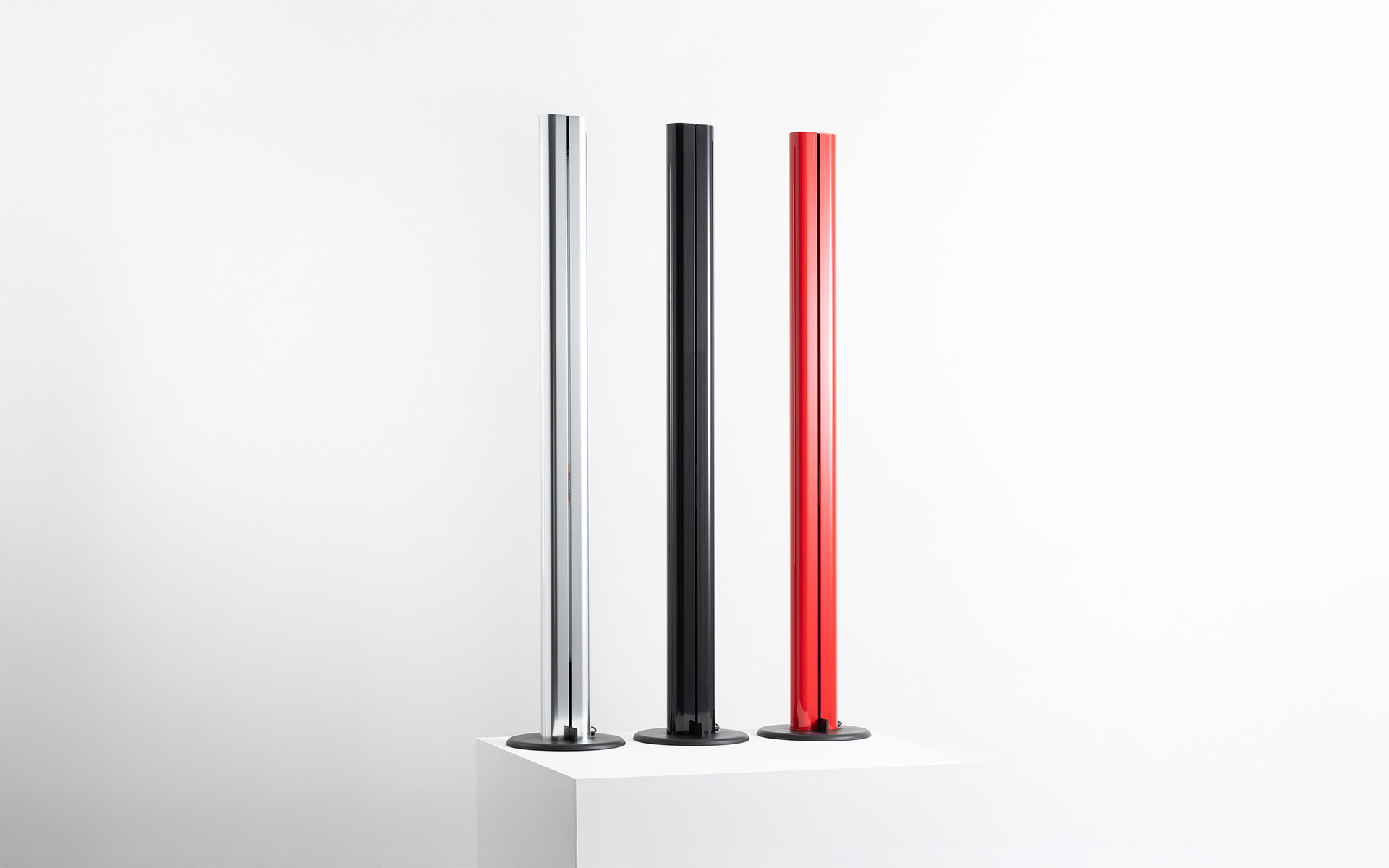 Tre lampade da terra sottili su basi rotonde in argento, nero e rosso, esposte su un piedistallo bianco su sfondo neutro.