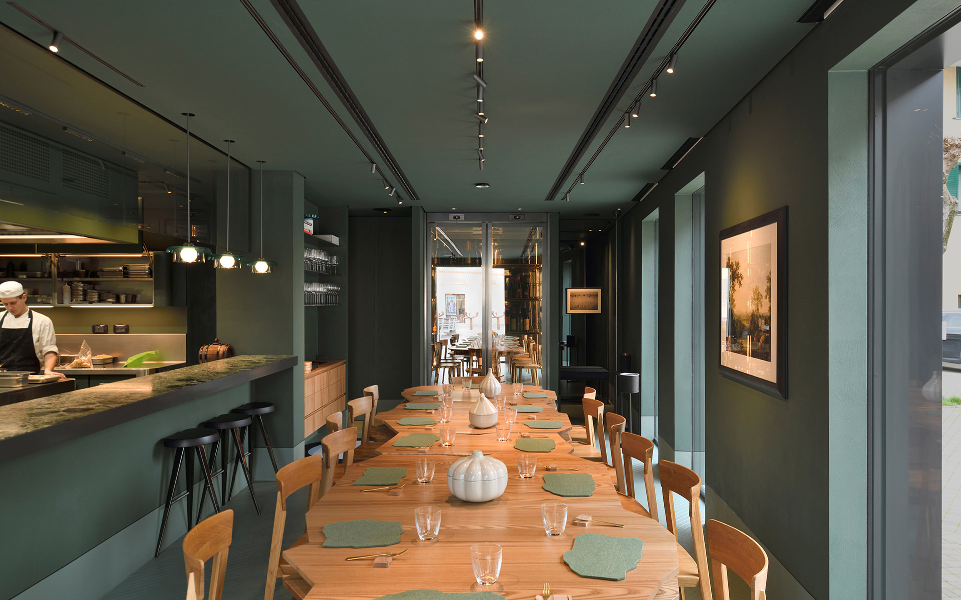 Sala ristorante con faretti Artemide su binario su soffitto verde petrolio, lungo tavolo in legno apparecchiato per cena, cucina a vista con sospensioni