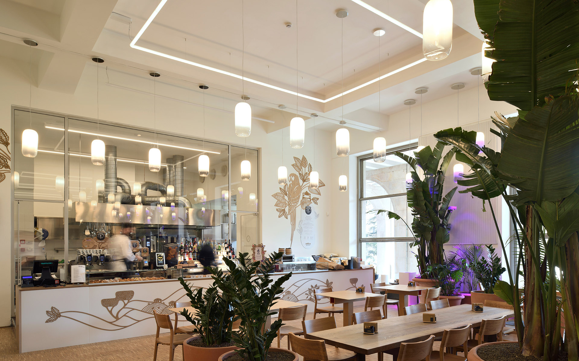 Interno di un moderno caffè con lampade a sospensione e applique Artemide, linee LED geometriche a soffitto, piante rigogliose e tavoli.
