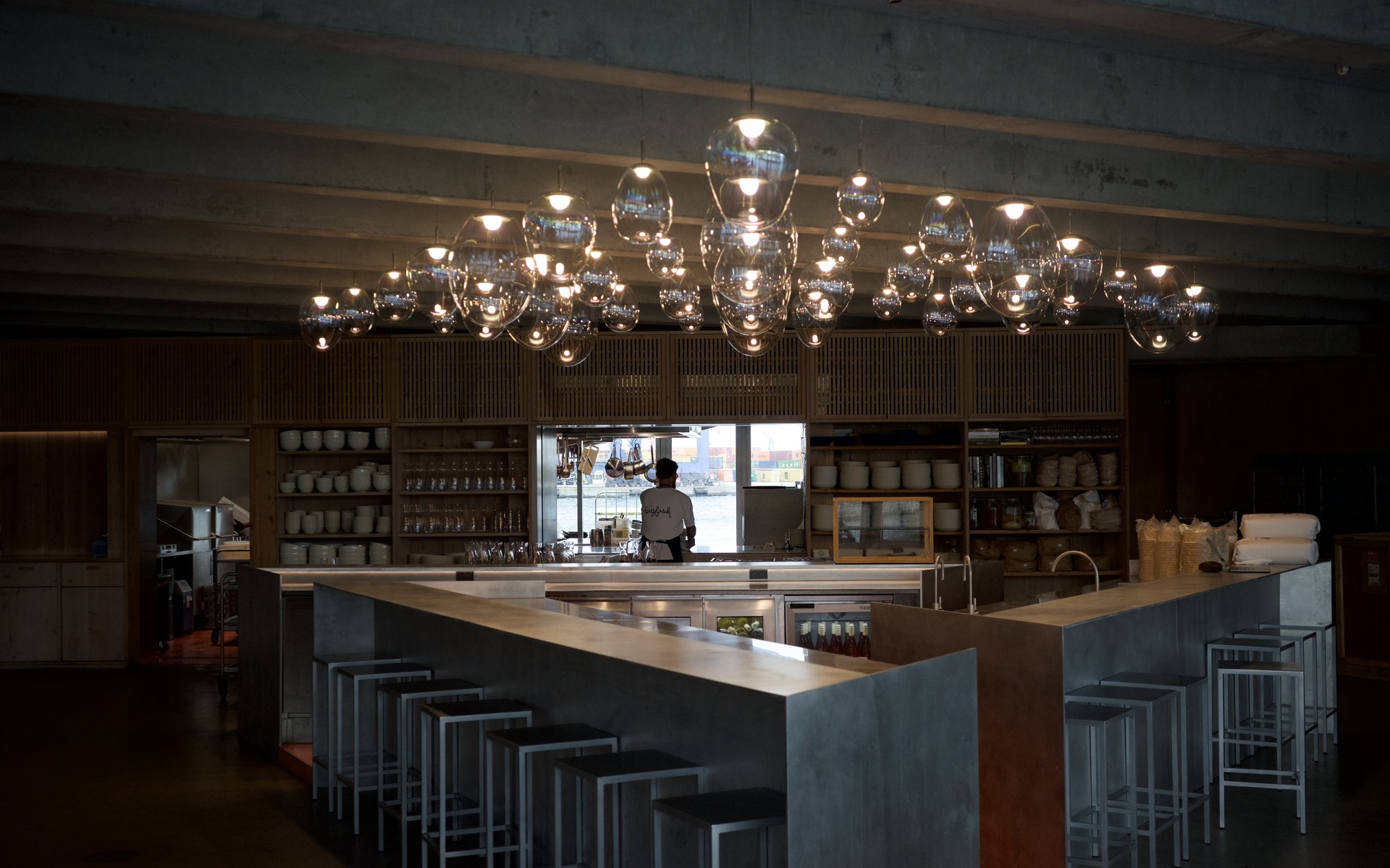 Bar ristorante con grande lampadario a grappolo di luci a sospensione in vetro che illumina un bancone in cemento con sgabelli metallici.