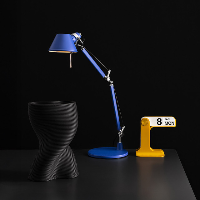 Lampada Tolomeo blu e calendario Timor giallo disposti su un tavolo con sfondo nero.