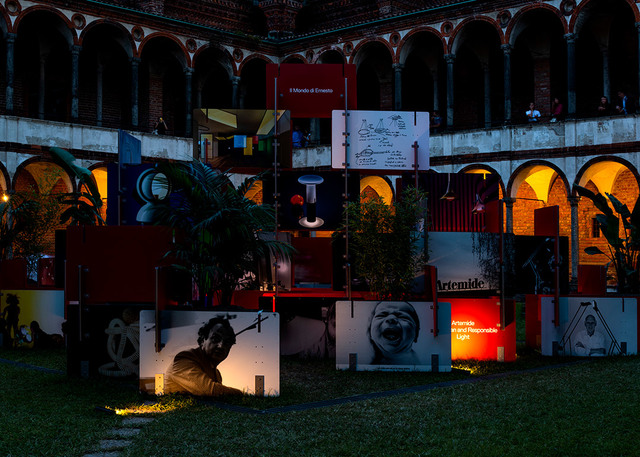 Artemide exhibition installation at night in a historic courtyard, featuring illuminated panels with brand imagery and lighting products on display.