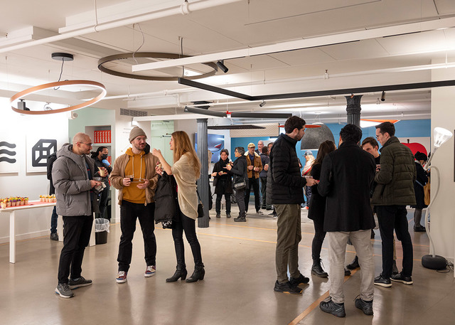 Gruppi di persone in networking a un evento interno, con lampade a sospensione circolari Artemide e faretti su binario che illuminano lo spazio.