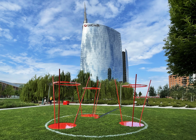 Red sculptural ring installations on green lawn in Porta Nuova district, Milan, with UniCredit Tower glass skyscraper in background.