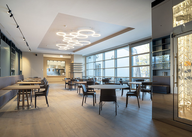 Interno di ristorante moderno con luci a sospensione LED circolari sul soffitto, faretti su binario sulla parete sinistra, grandi finestre e mobili in legno scuro.