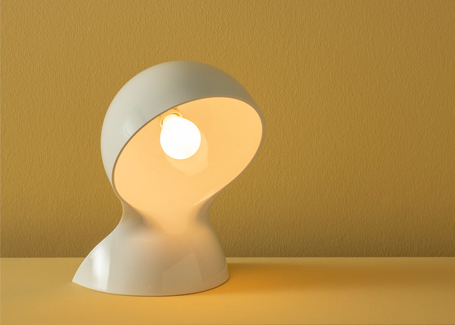 Artemide Dalù white table lamp with sculptural tilted shade revealing a warm glowing bulb, casting soft light on a yellow surface