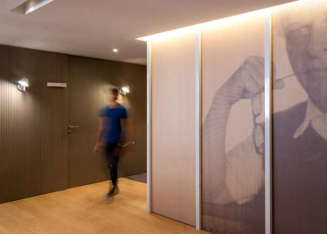 Corridoio moderno con illuminazione a LED calda a soffitto, faretti a parete e una persona che cammina davanti a un pannello decorativo stampato.