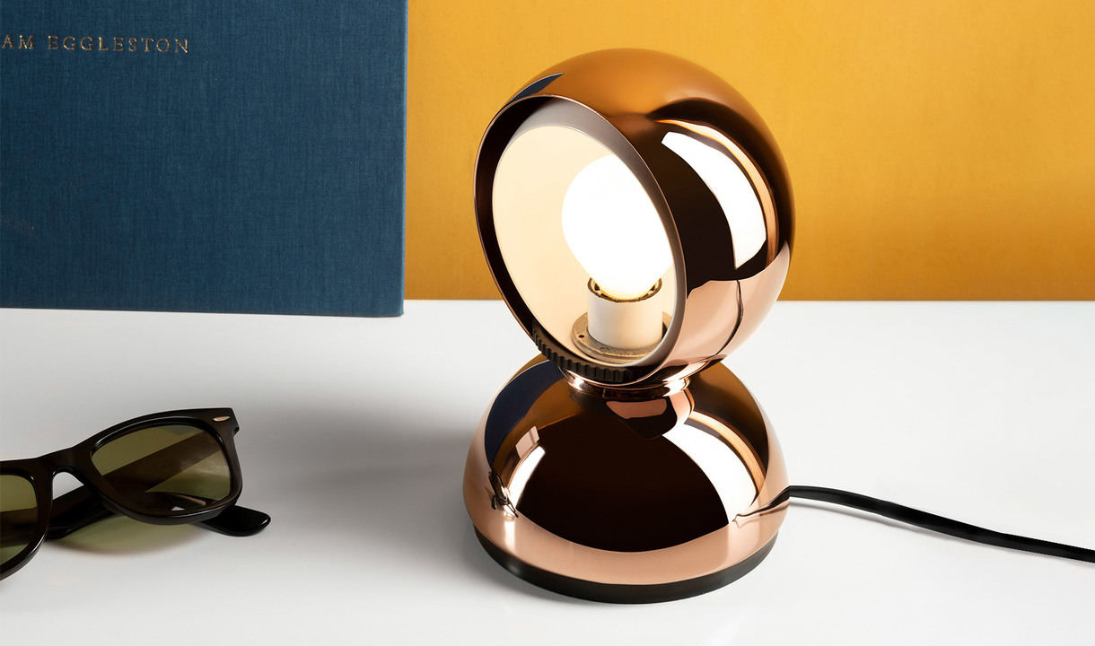Artemide Eclisse table lamp in copper finish, spherical rotating shade revealing bulb, placed on white desk with sunglasses and book