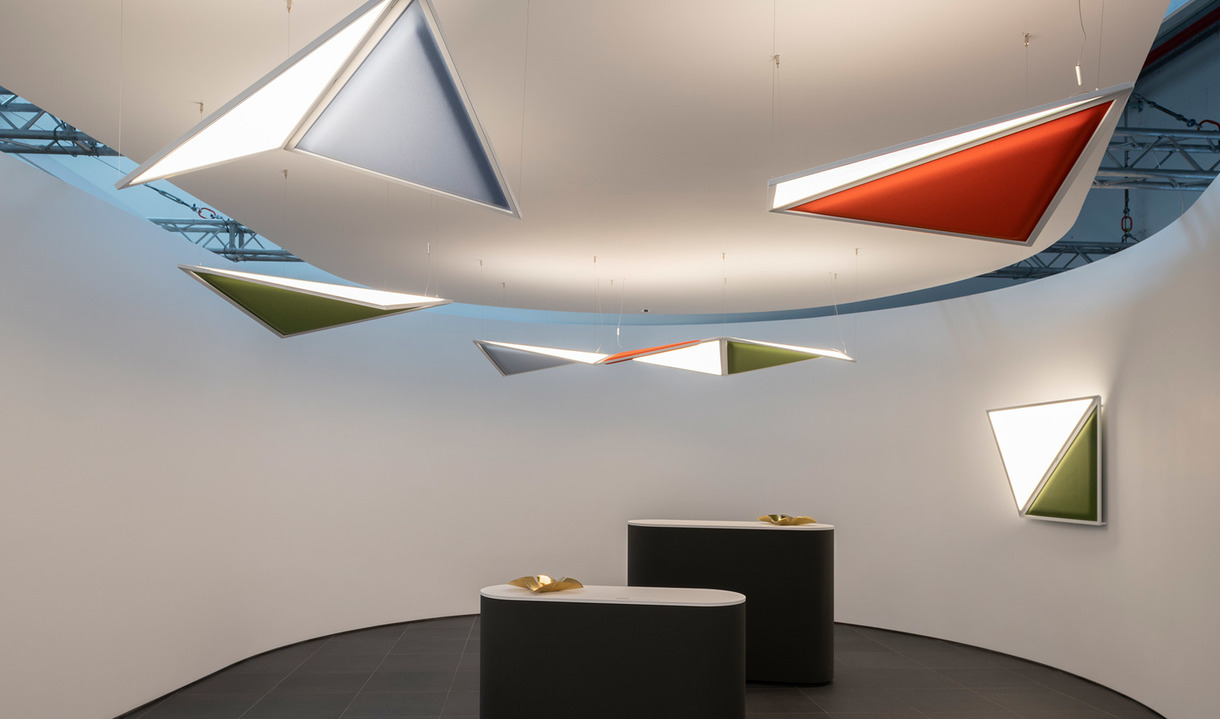 Lampade a sospensione e da parete triangolari in bianco, rosso, verde e blu in una sala curva bianca sopra due banconi scuri.