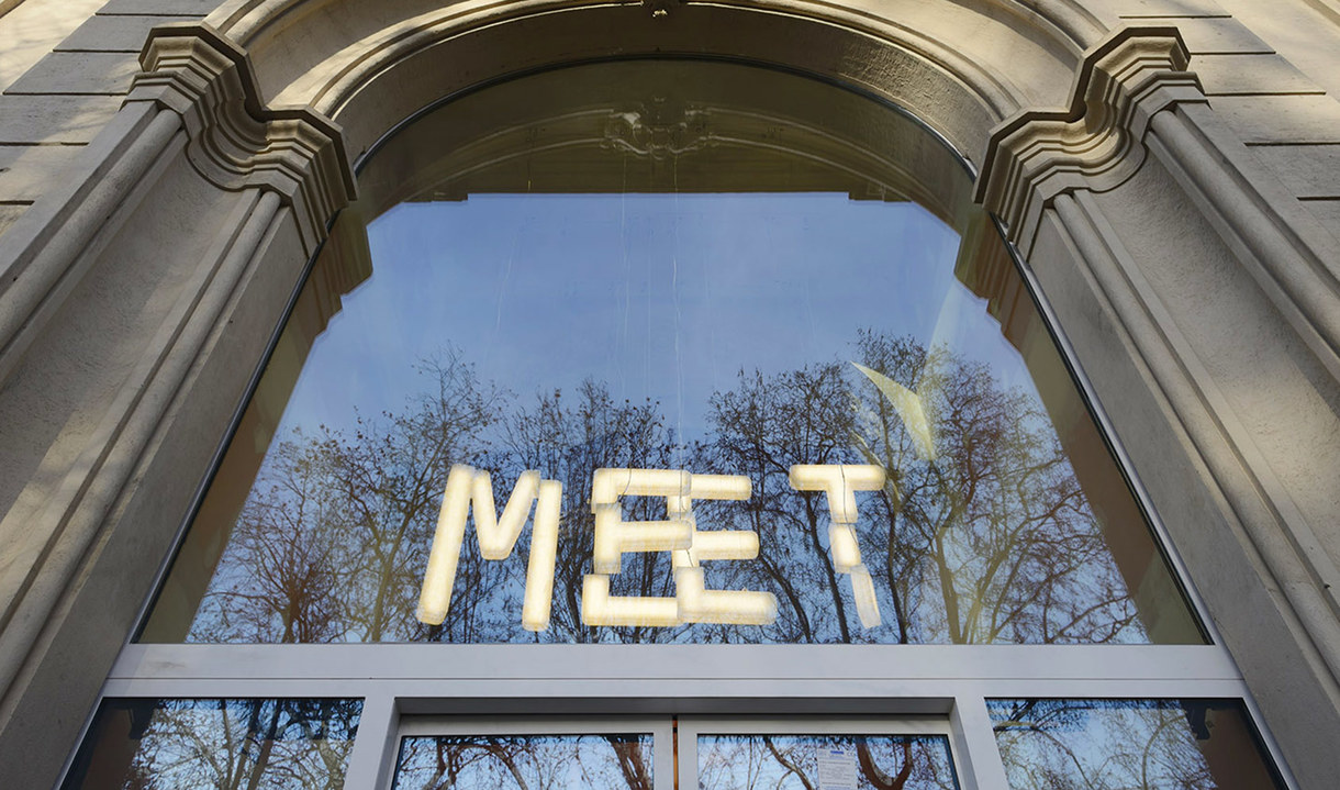 Illuminated 'MEET' letters made of tubular lights displayed in a large arched window of a classical stone building.