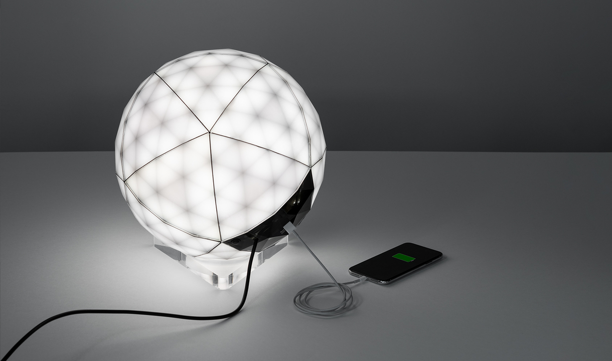 Lampada da tavolo LED sferica Artemide con diffusore sfaccettato geometrico, su base trasparente, con porta USB e smartphone collegato