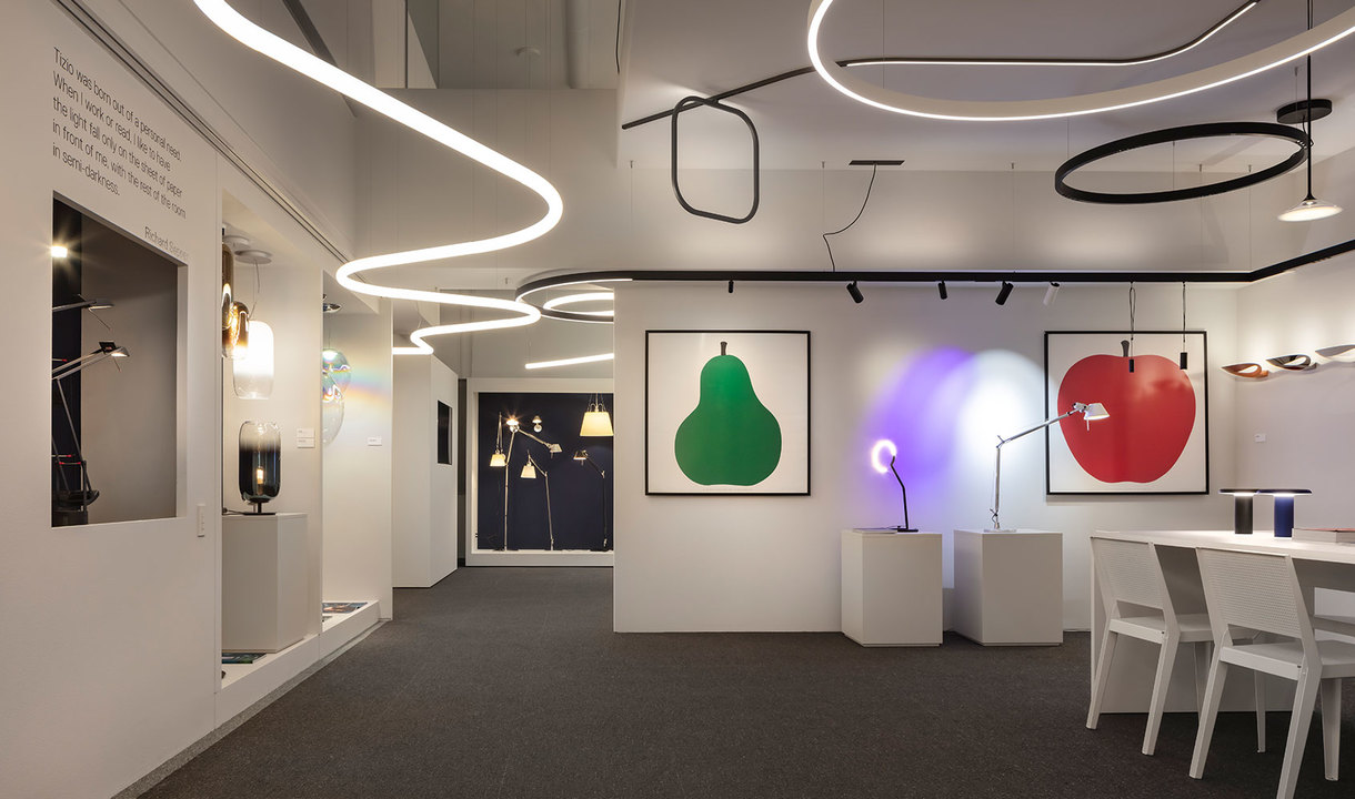 Artemide showroom featuring sinuous LED ceiling lights, circular pendant lamps, floor lamps, and desk lamps displayed on white pedestals and tables