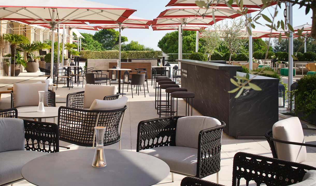 Elegante terrazza esterna con sedie lounge in intreccio nero, tavolini rotondi con lampada da tavolo sottile, grandi ombrelloni e bancone bar.