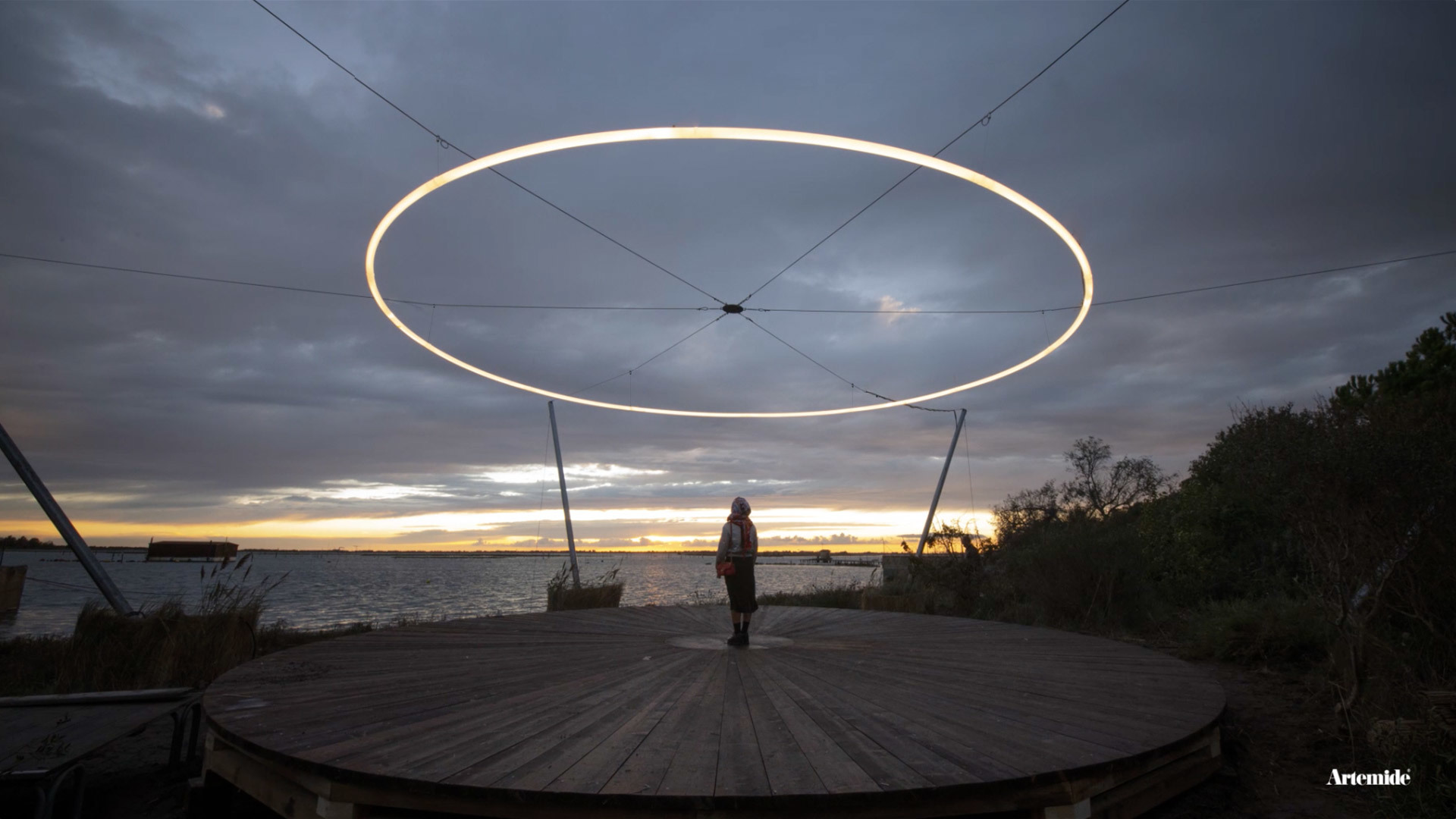 Persona su una terrazza in legno sotto un grande anello luminoso circolare Artemide sospeso all'aperto al tramonto sul lungomare.