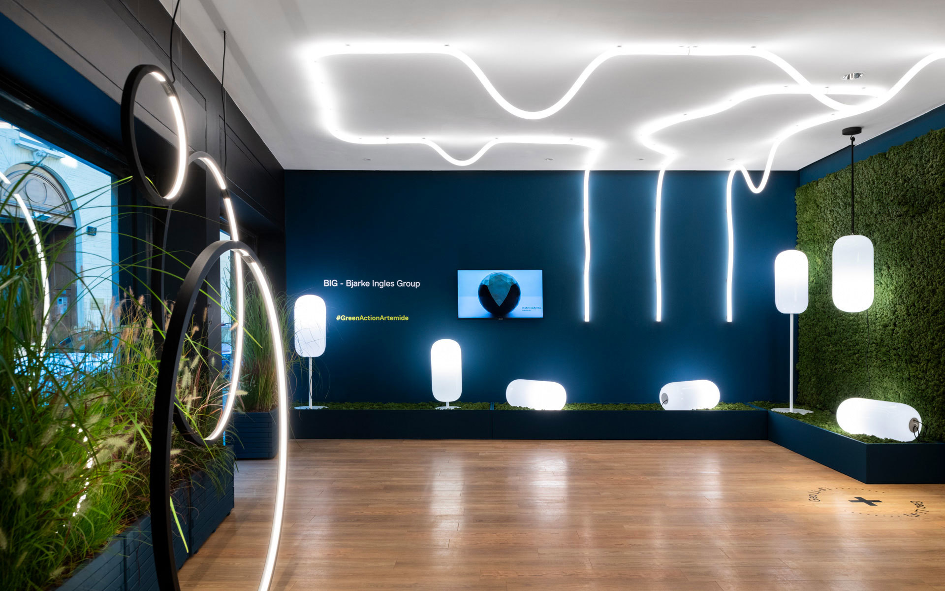 Showroom Artemide con collezione BIG: linee LED organiche a soffitto, lampade da terra circolari e sospensioni ovali bianche su parete verde acqua con piante.