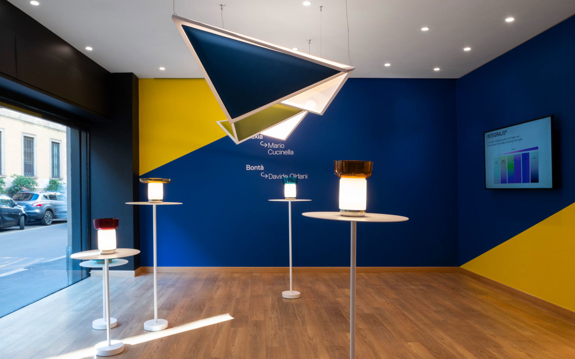 Showroom Artemide con lampada a sospensione geometrica e lampade da tavolo Bontà di Davide Groppi su steli, su parete blu e gialla