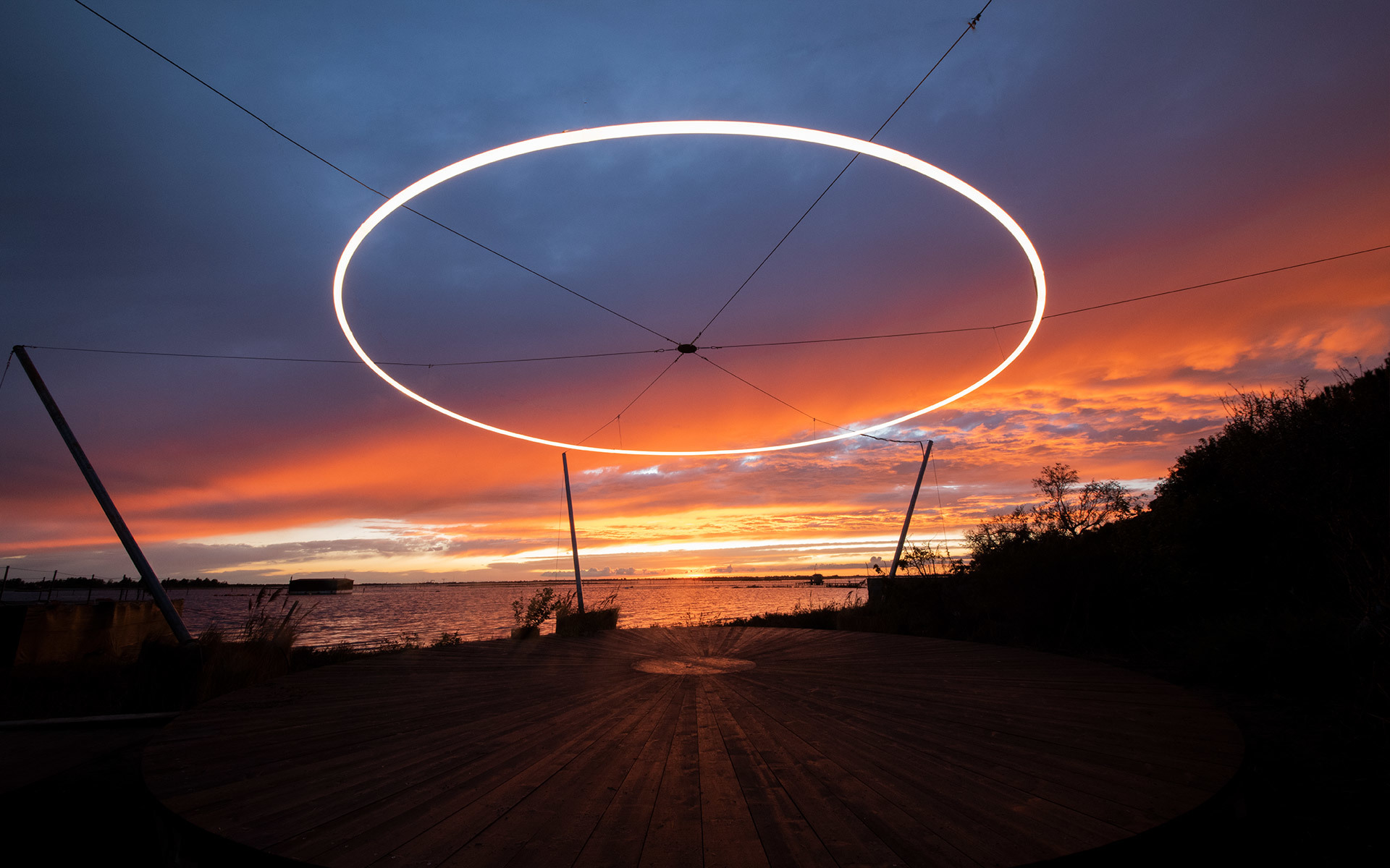 Grande anello luminoso bianco sospeso all'aperto su un cielo al tramonto rosso e arancione sull'acqua, sorretto da cavi