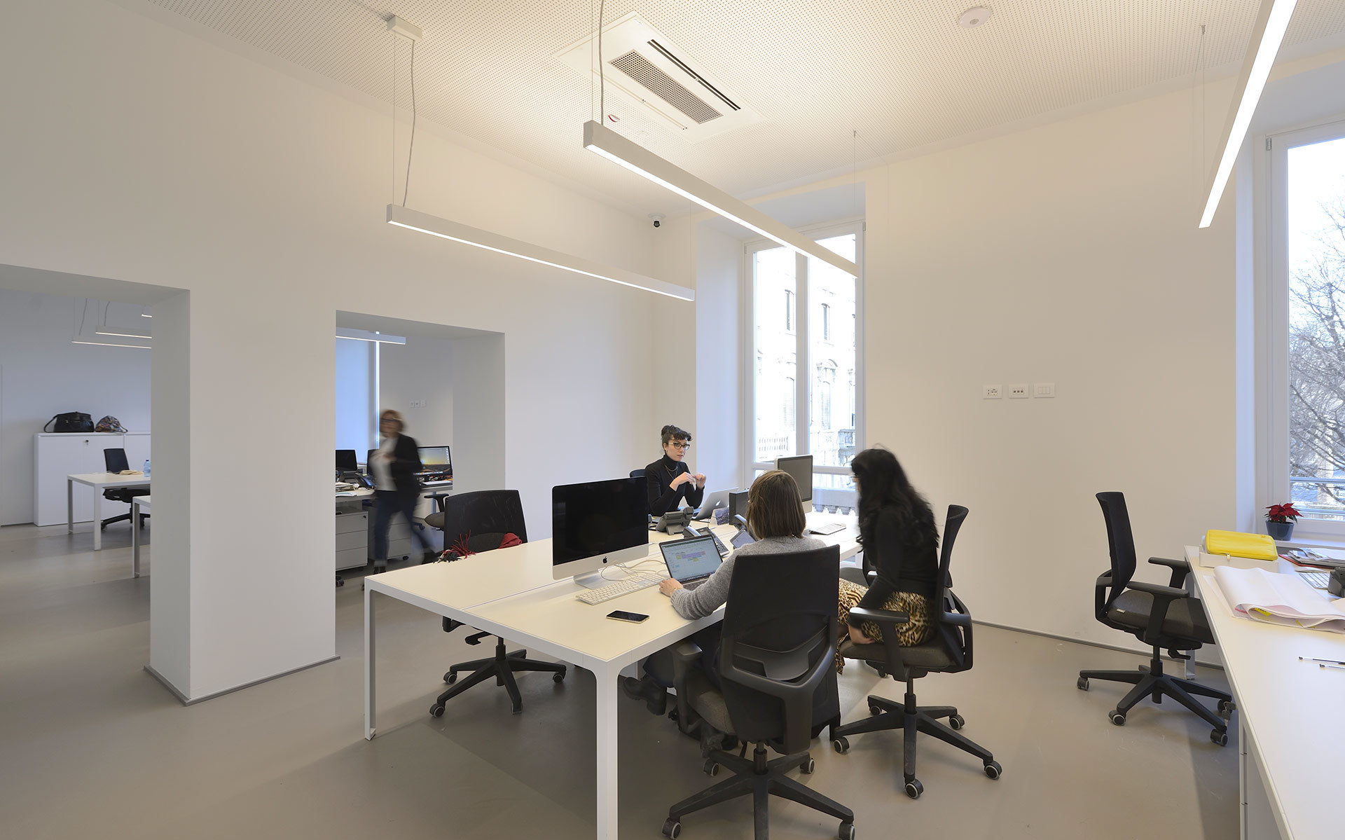 Modern office with white walls, workers at desks with monitors, lit by Artemide linear pendant and recessed ceiling lights.