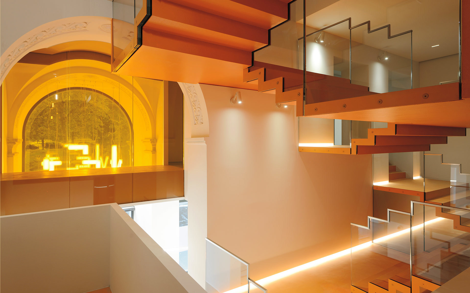 Modern staircase with glass railings and warm LED strip lighting under steps, illuminating an elegant interior with an arched window.
