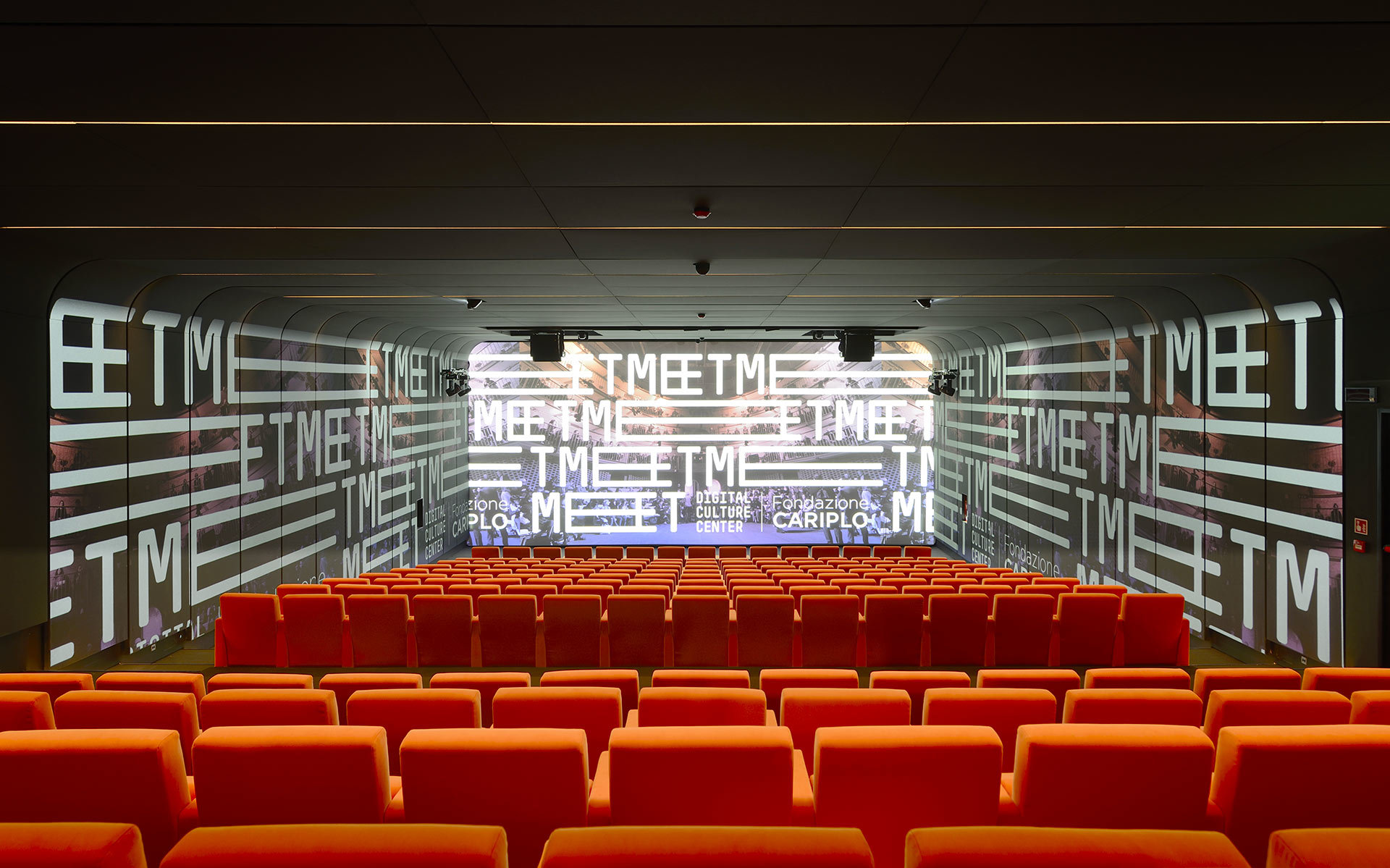 Cinema auditorium with red seats facing a wide projection screen displaying 'MEET' text, with ceiling-mounted spotlights illuminating the space.