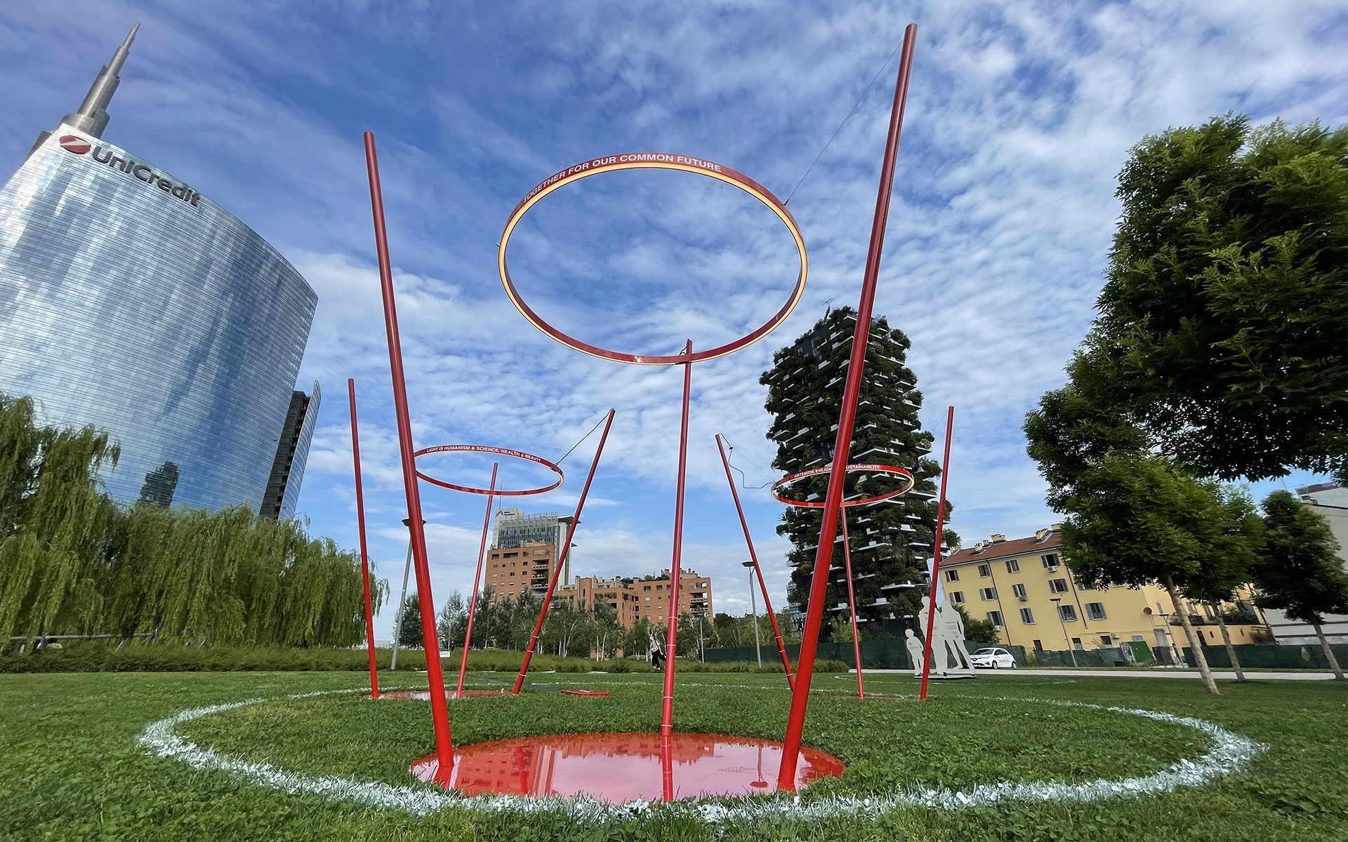 Red sculptural installation with tall poles and circular rings on green lawn, with UniCredit Tower and Bosco Verticale in Milan background