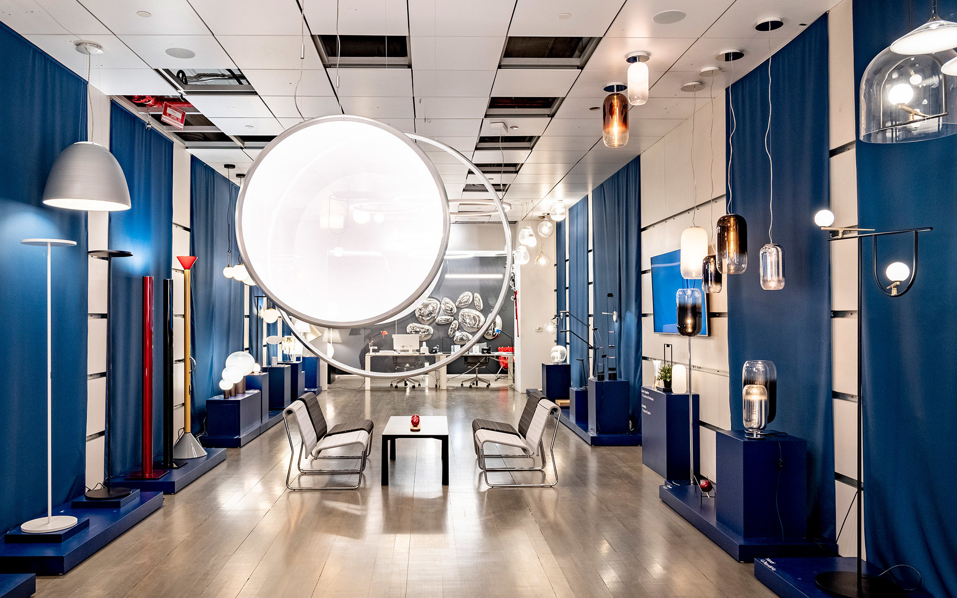 Showroom Artemide con lampade a sospensione, da terra e da parete esposte lungo pareti con tende blu, al centro una grande installazione luminosa circolare.