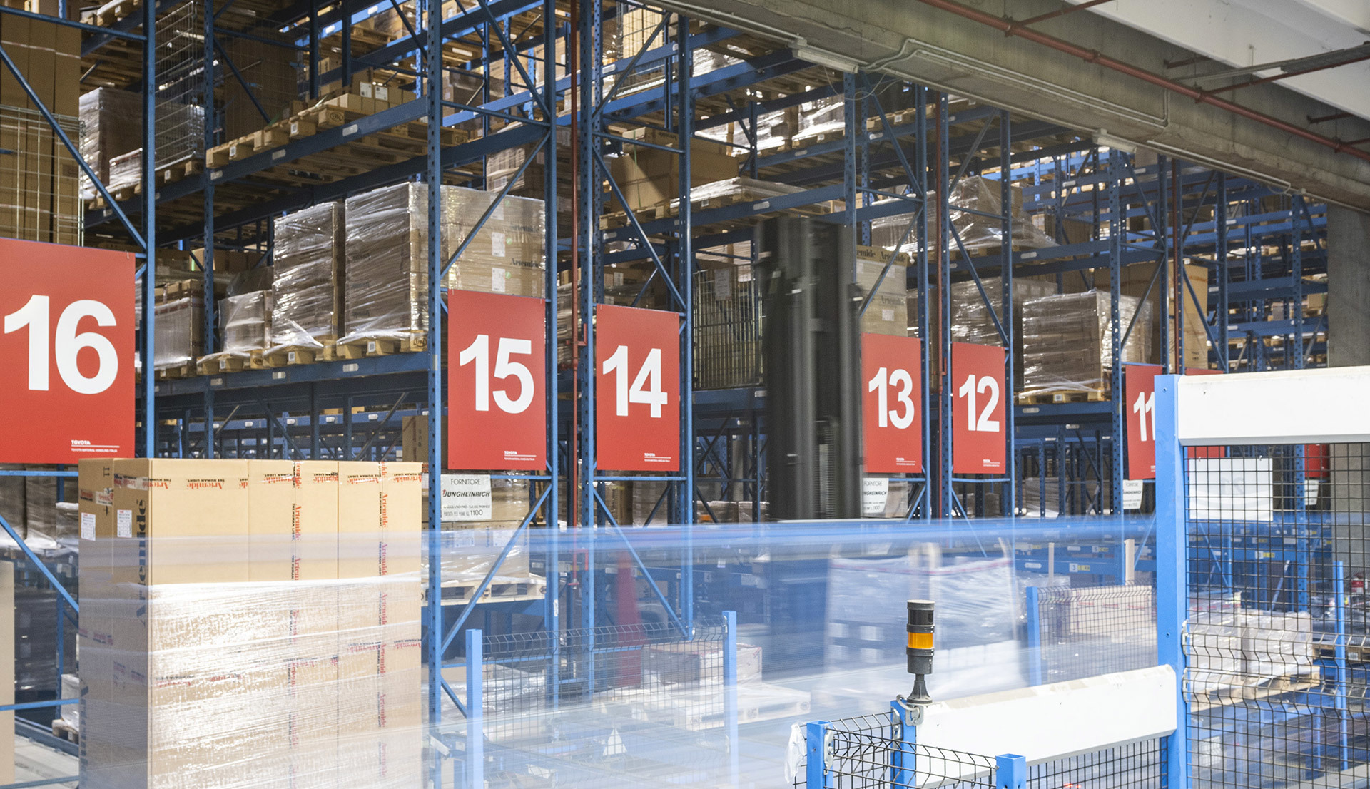 Interno di un grande magazzino con scaffalature metalliche blu numerate (11-16) che stoccano merci pallettizzate su più livelli.