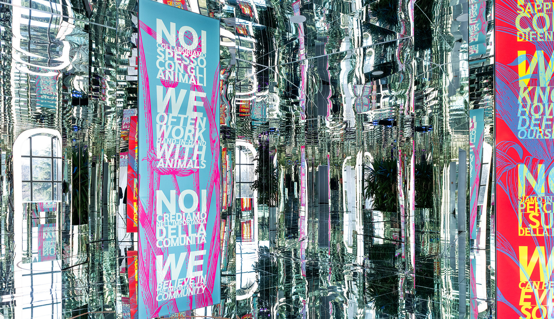 Installazione immersiva con specchi e striscioni colorati appesi con testi bilingue su comunità e animali