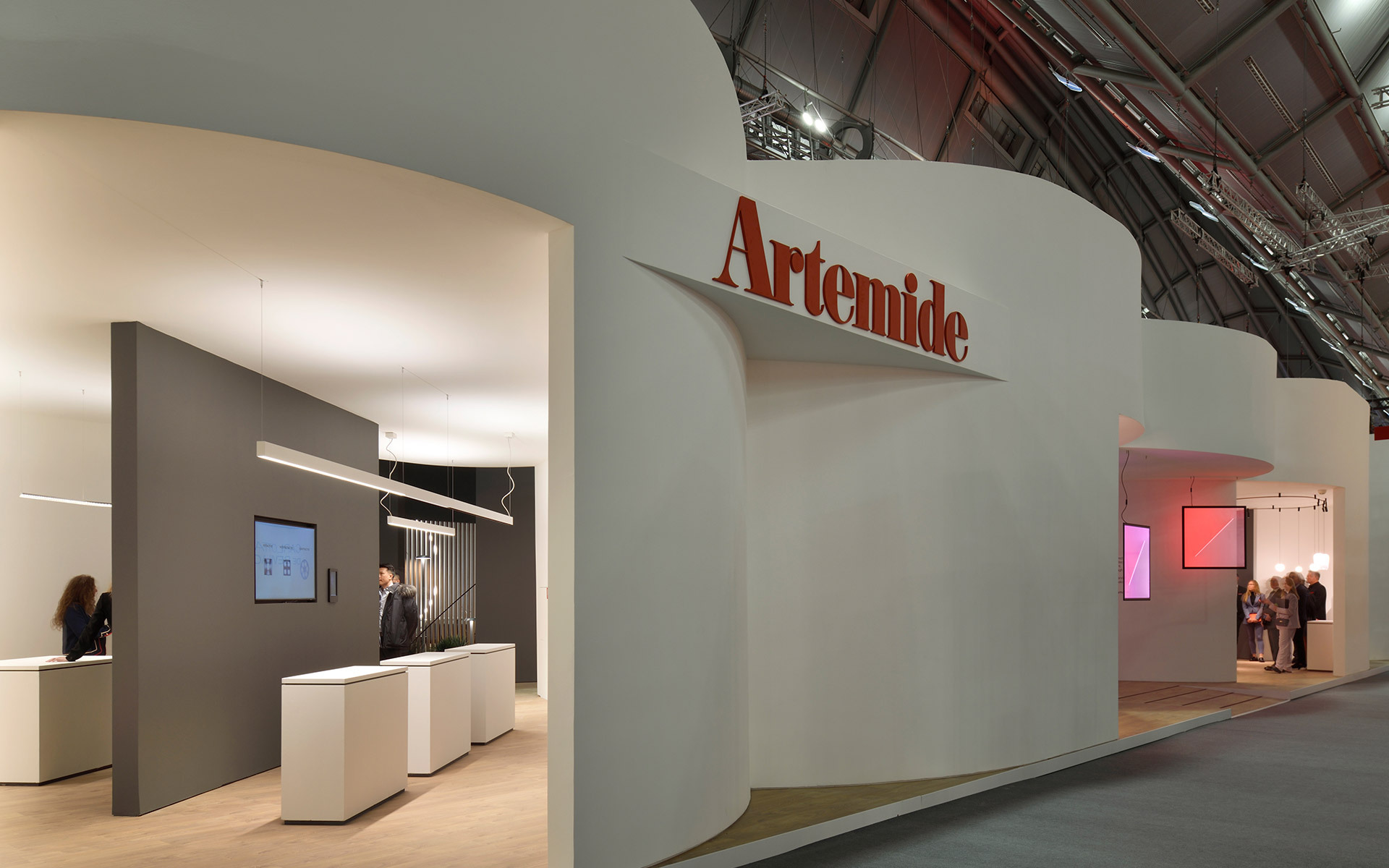 Stand espositivo Artemide con pareti curve bianche, logo, e uno showroom interno scuro con lampade a sospensione e lineari.