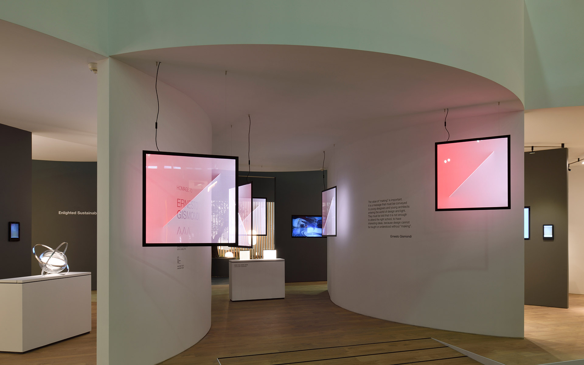 Spazio espositivo moderno con pareti bianche curve, schermi digitali sospesi con immagini rosa e una lampada scultorea bianca su piedistallo.