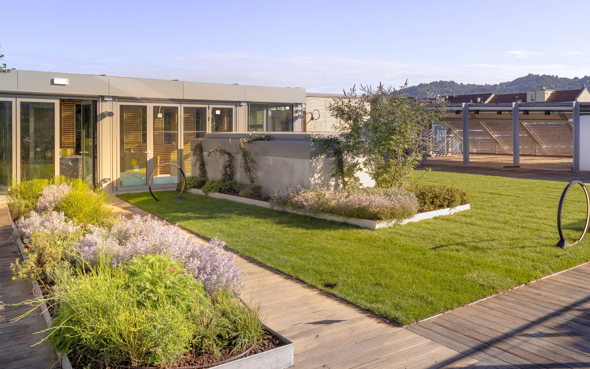 Giardino pensile con prato verde rigoglioso, piante fiorite in fioriere di legno e un edificio moderno con ampie vetrate.