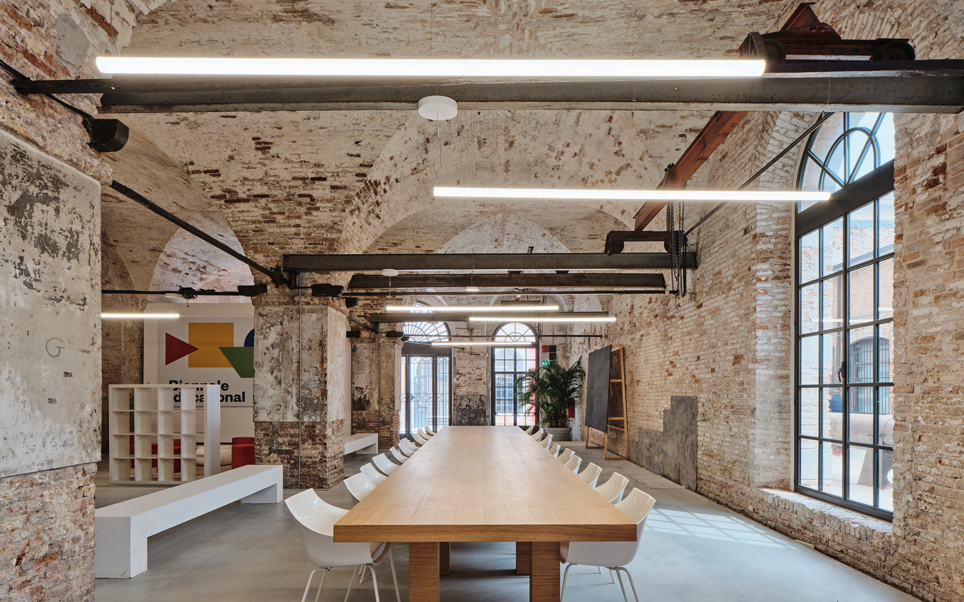 Conference room in historic brick-vaulted space with long wooden table, white chairs, and linear pendant lights illuminating the interior.