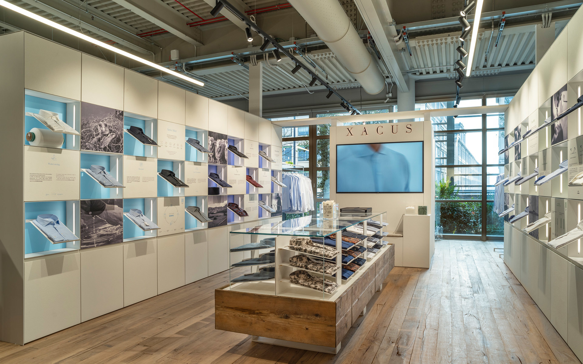 Interno del negozio Xacus con lampade lineari LED Artemide a soffitto che illuminano le pareti espositive e il tavolo centrale in legno.