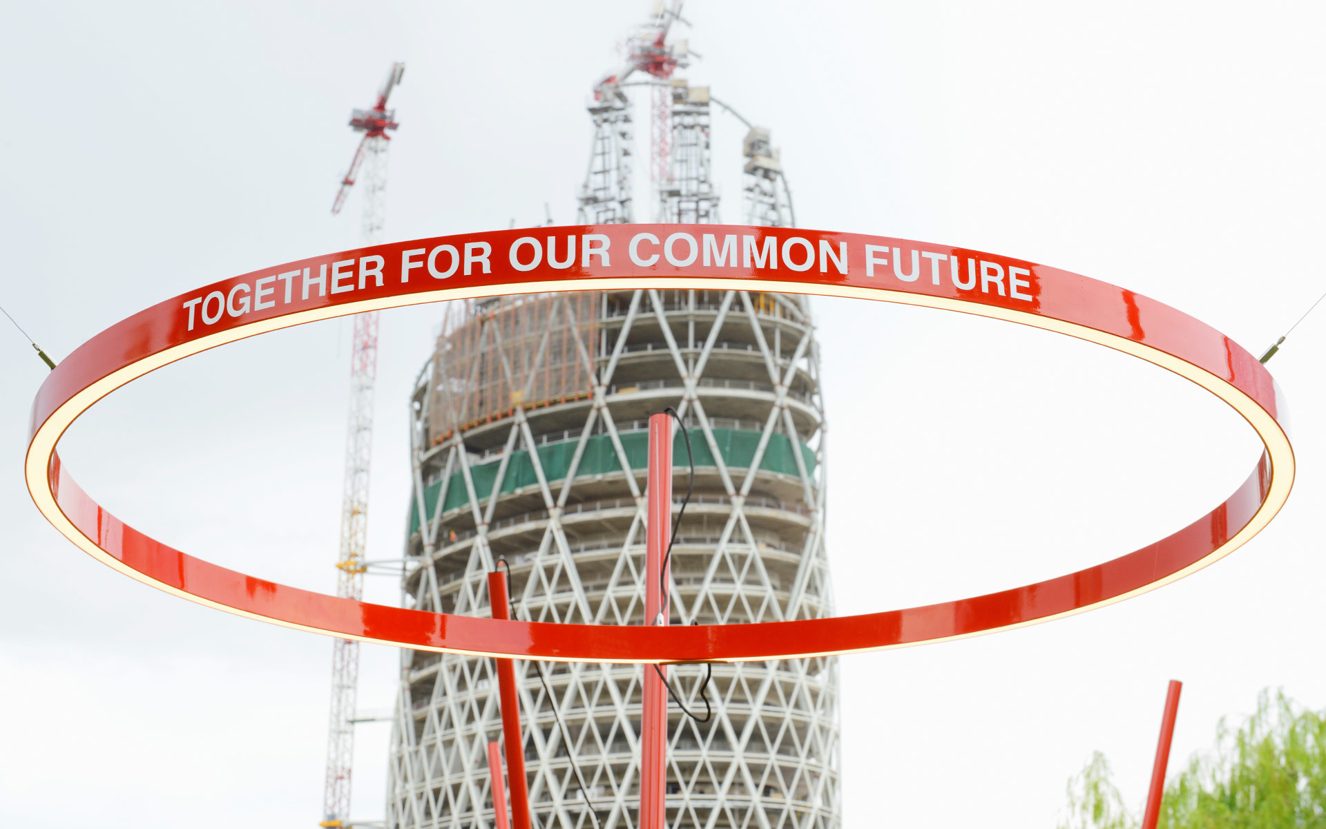 Large red illuminated ring with 'Together For Our Common Future' text, suspended before a tower under construction with cranes