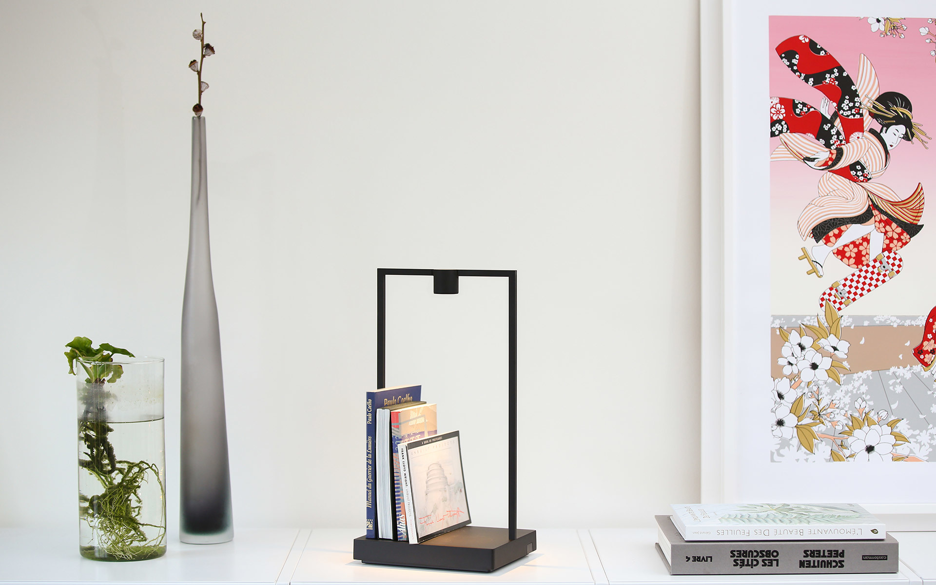 Lampada da tavolo con struttura rettangolare nera e base in legno che regge piccoli libri, su mensola bianca con vasi decorativi e opera d'arte.