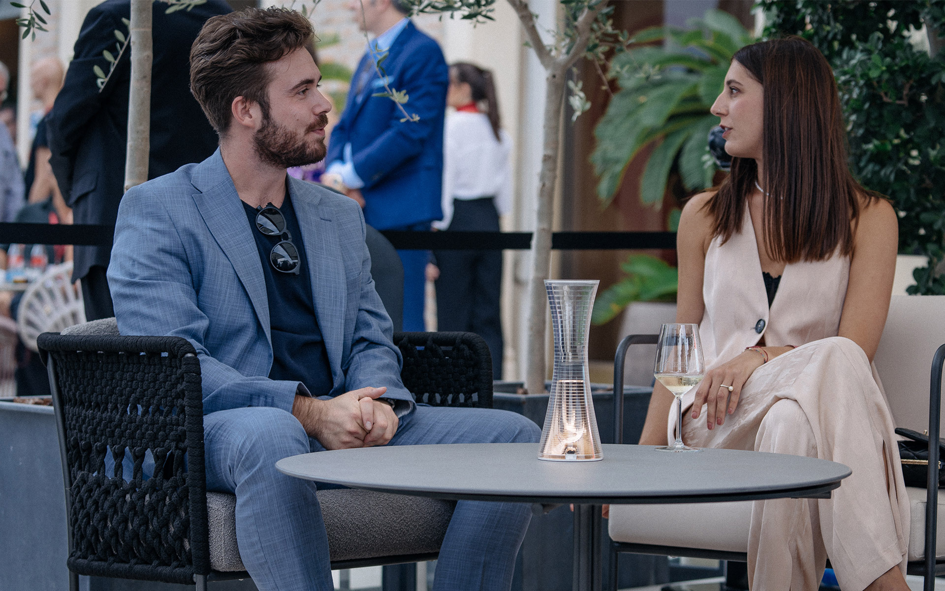 Due persone in conversazione a un tavolo di caffè all'aperto con una lanterna in vetro Artemide al centro e un calice di vino.