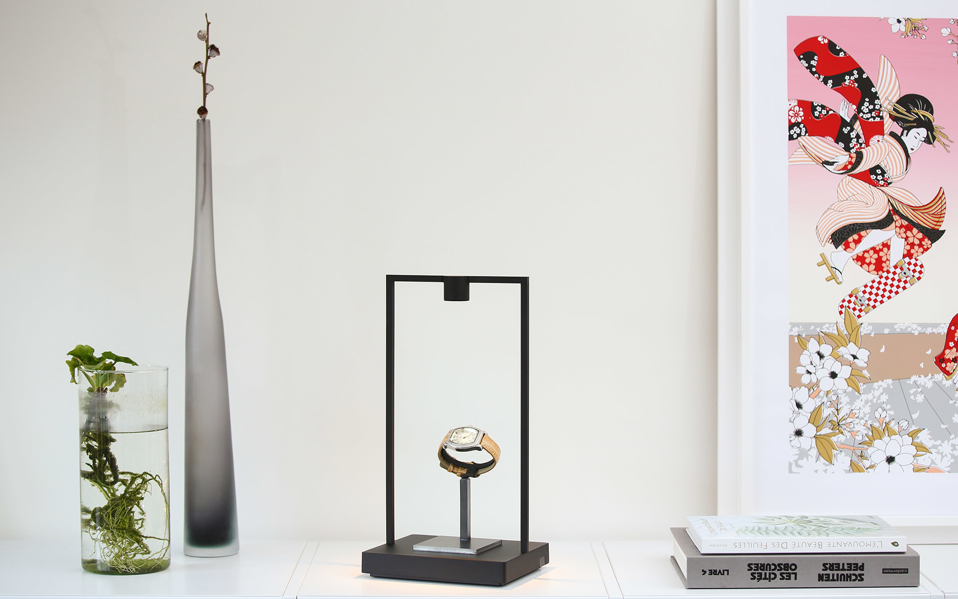 Lampada da tavolo con cornice rettangolare nera che espone un orologio, stilizzata come supporto espositivo minimalista su mensola bianca con oggetti decorativi