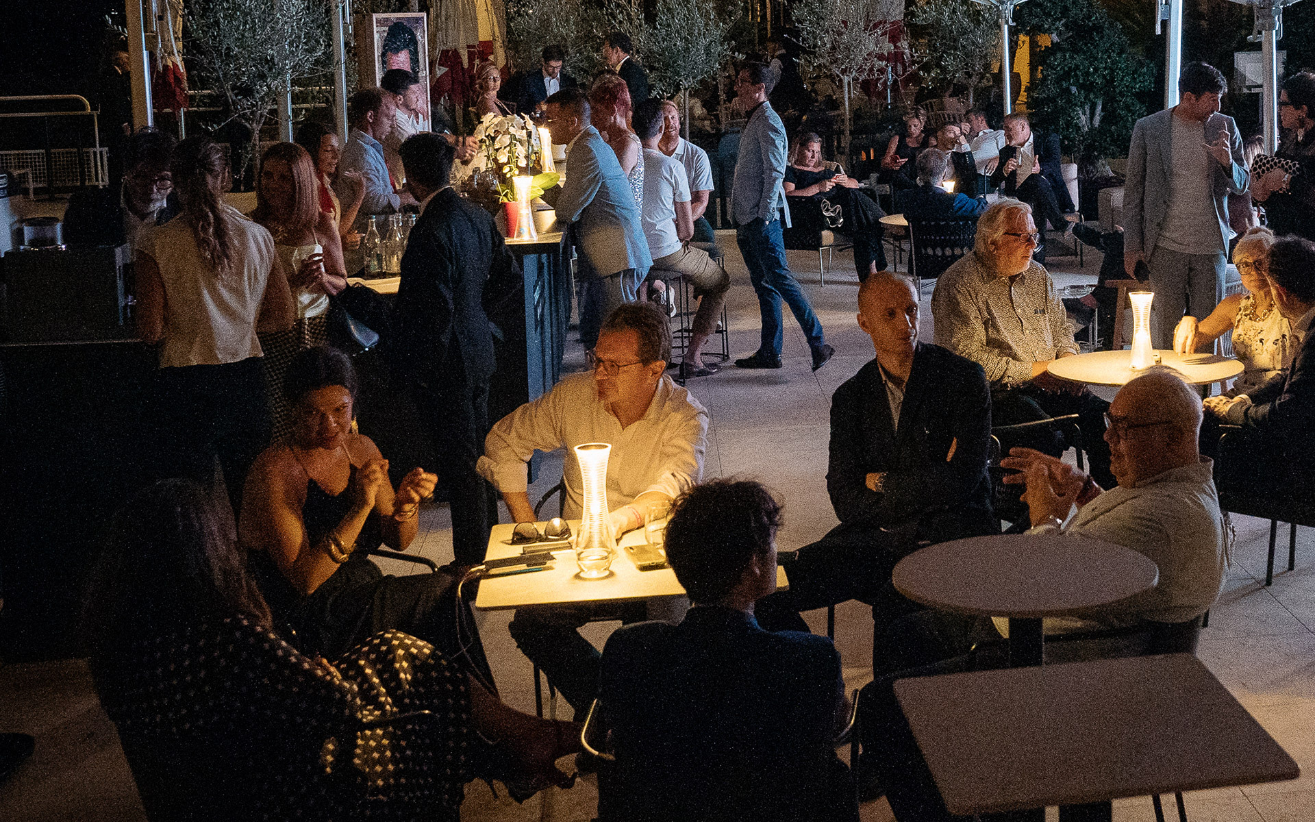 Serata all'aperto con persone che socializzano ai tavoli, illuminate da una lampada da tavolo portatile con luce calda al centro.