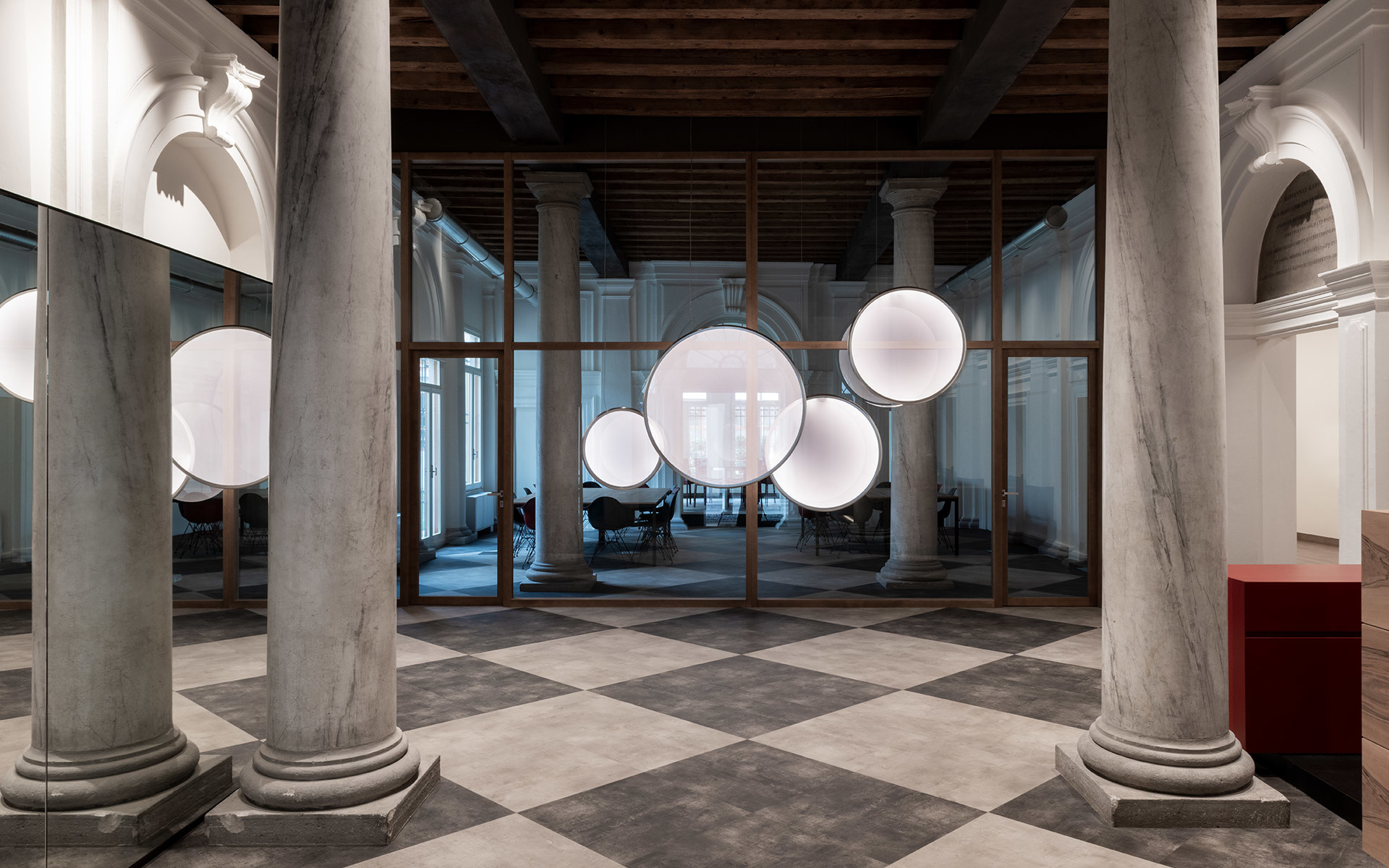 Gruppo di lampade a sospensione sferiche luminose in una sala storica con colonne in marmo e pavimento a scacchi.