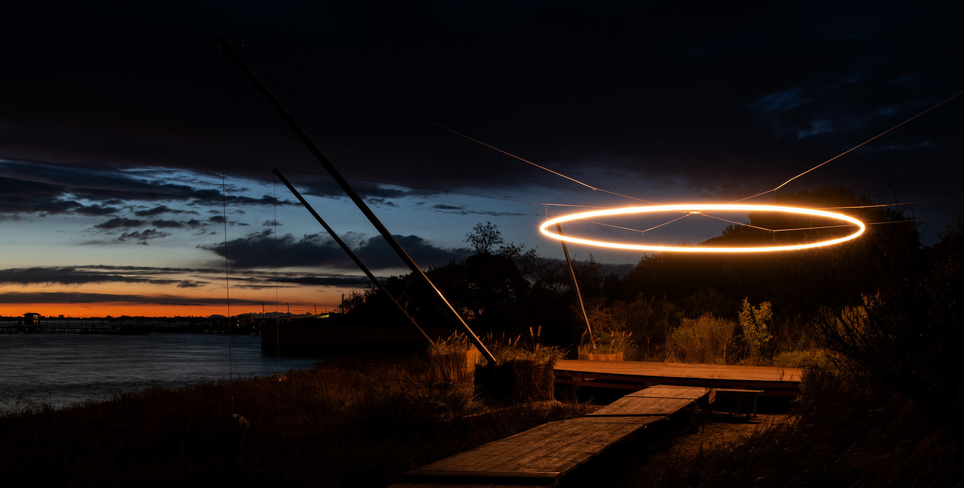 Installazione luminosa ad anello LED circolare sospesa all'aperto al crepuscolo, riflessa sul lungofiume con cielo al tramonto