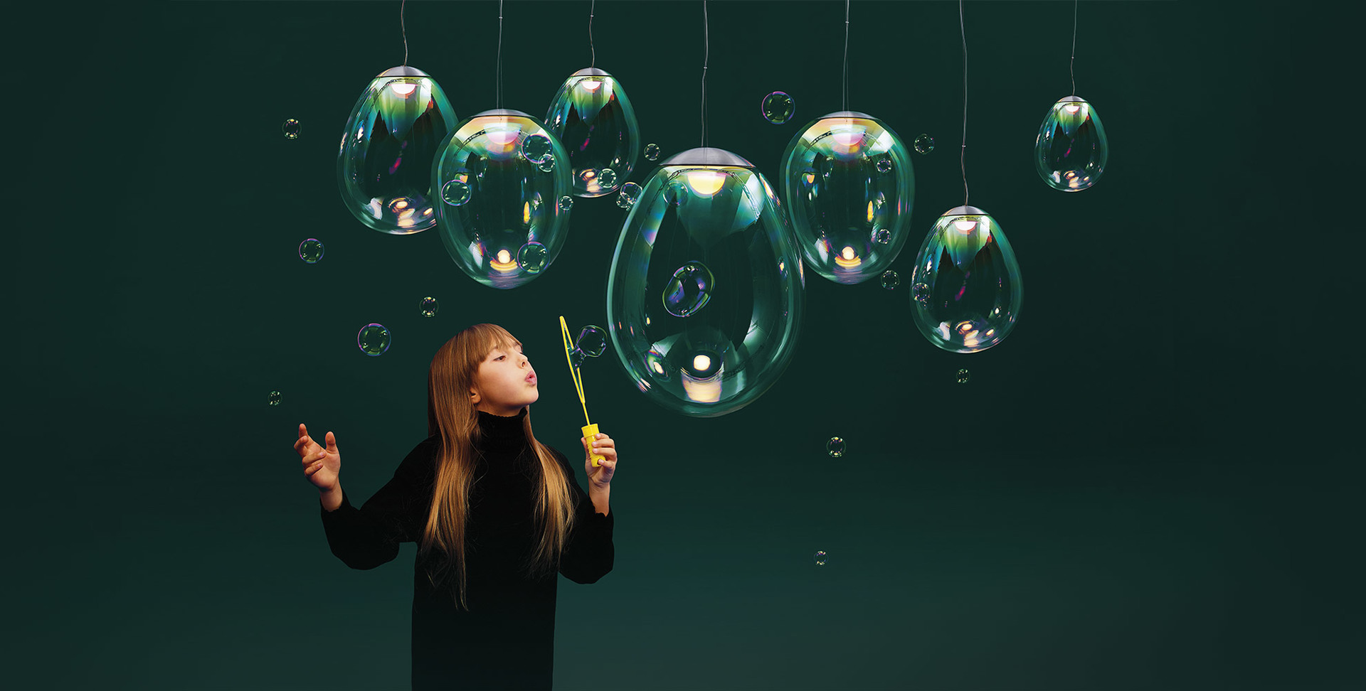 Bambina che soffia bolle di sapone vicino a lampade a sospensione Artemide in vetro iridescente simili a bolle fluttuanti su sfondo verde scuro