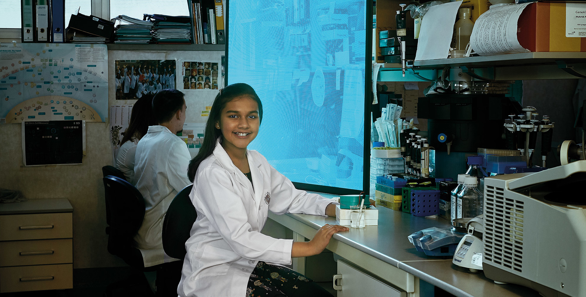 Giovane scienziata in camice bianco sorridente alla fotocamera in un laboratorio, collega al computer sullo sfondo