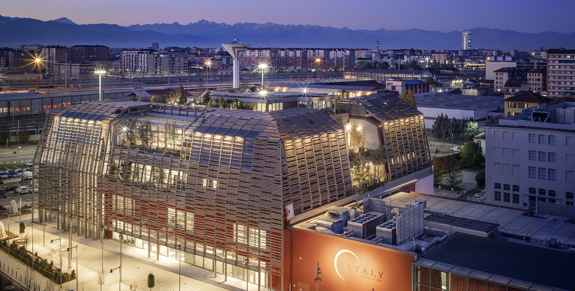 Vista aerea al crepuscolo di Eataly Torino, edificio moderno con facciata a reticolo in legno illuminata e calda luce interna sullo skyline alpino