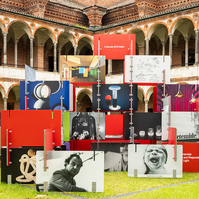 Artemide exhibition in a historic arcaded courtyard featuring panels with lamp designs, sketches, and portraits, titled 'Il Mondo di Ernesto'