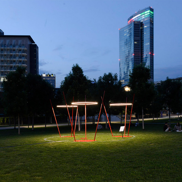 Artemide luminous ring installations on red angular supports illuminate a park at dusk, with city buildings in the background.