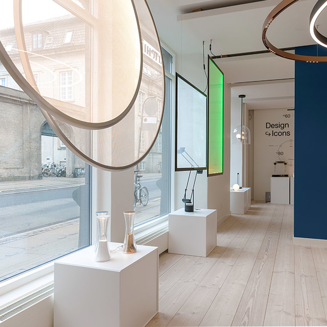 Showroom Artemide con collezione Design Icons: anelli circolari a sospensione, lampade da tavolo su plinti bianchi, pannelli retroilluminati colorati.